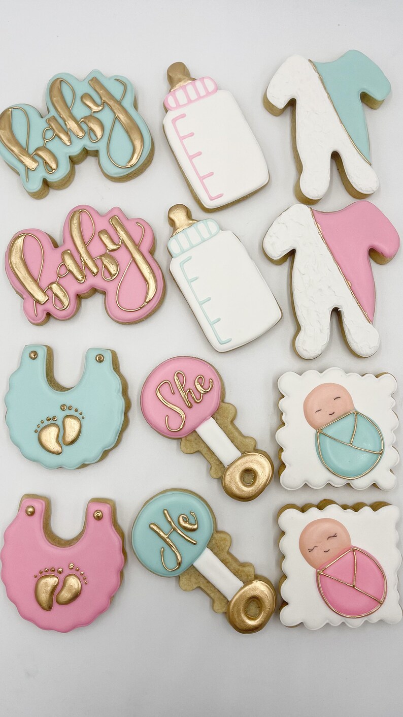 K&ouml;nnte beinhalten: Ein Set aus zehn dekorierten Zuckerkeksen mit Baby-Motiv. Die Kekse sind mit rosa, blauem und goldenem Zuckerguss verziert. Die Kekse zeigen die W&ouml;rter "Baby", "Sie", "Er" und Bilder von Babyflaschen, Stramplern und Rasseln.
