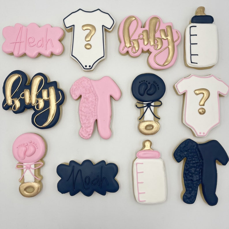K&ouml;nnte beinhalten: Zw&ouml;lf dekorierte Zuckerkekse mit verschiedenen Designs, darunter Baby-Bodys, eine Babyflasche, eine Rassel und die Worte "Baby" und "Noah". Die Kekse sind mit rosa, blauem und goldenem Zuckerguss verziert.
