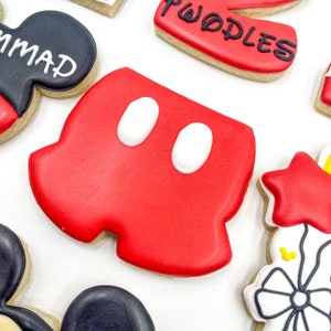May include: Red and white cookie decorated with Mickey Mouse's iconic shorts. The cookie is shaped like a pair of shorts with two white circles on the front.
