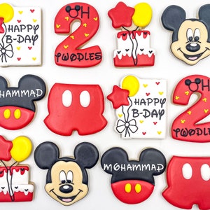 Op de afbeelding: Een assortiment van tien gedecoreerde suikerkoekjes met Mickey Mouse, een verjaardagstaart en de cijfers 2 en 8. De koekjes zijn versierd met rode, witte, zwarte en gele glazuur. De tekst op de koekjes luidt "Happy B-Day", "Mohammed", "Twoodles" en "Happy B-Day".