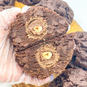 May include: Close-up of a chocolate chip cookie with a hazelnut chocolate center. The cookie is broken in half, revealing the filling. The cookie is on a wooden surface.