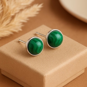 May include: A pair of stud earrings featuring round, polished malachite stones set in silver. The earrings display a vibrant green color with unique swirling patterns. The earrings are presented on a brown box, suggesting a jewelry display.