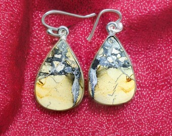 May include: A pair of teardrop-shaped earrings with a silver-colored metal setting. The earrings feature a natural stone with a mix of beige, black, and gray patterns. The earrings are displayed against a red background.
