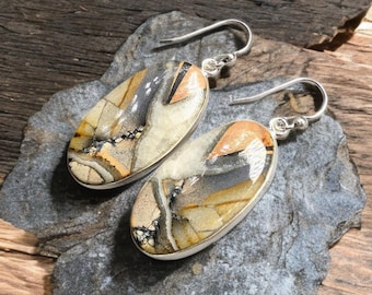 May include: Oval-shaped earrings with a silver frame. The earrings feature a unique pattern of beige, brown, black, and orange hues. The earrings are displayed on a gray stone surface with a wooden background.