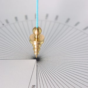 Può includere: Un pendolo in ottone con uno spago blu appeso sopra un goniometro circolare bianco con linee e numeri neri. Il pendolo è appeso sopra il numero 0.