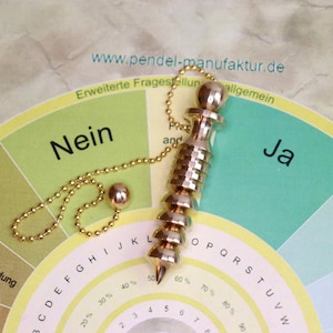 Puede incluir: Un péndulo dorado con una cadena y un peso redondo, apoyado en un gráfico circular. El gráfico tiene segmentos con texto, incluyendo "Nein" y "Ja". El sitio web "www.pendel-manufaktur.de" es visible.
