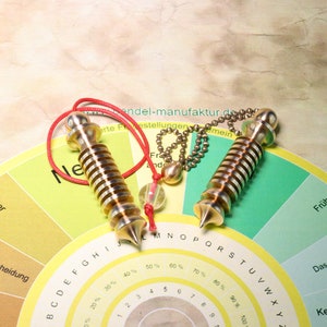 Puede incluir: Dos varillas de péndulo de radiestesia de color dorado con cuerdas rojas y una cadena. Las varillas están sobre un gráfico circular con las palabras "Später", "Deine Entscheidung", "nicht", "Früher", "Das entscheidest" y "Keine Antwort" impresas en él.