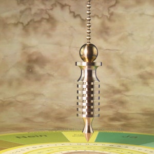 Puede incluir: Un péndulo de color dorado con una punta puntiaguda cuelga sobre un gráfico circular con las palabras "Nein" y "Ja" impresas en él. El gráfico está dividido en secciones de diferentes colores y números.