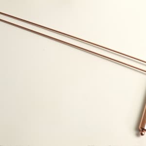 Op de afbeelding: Twee lange, dunne koperen staven met afgeronde uiteinden. De staven zijn in de buurt van de uiteinden gebogen in een hoek van 90 graden, waardoor een haakvorm ontstaat.