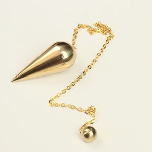 Puede incluir: Un péndulo dorado con un peso en forma de lágrima y una pequeña bola al final de la cadena. La cadena y los pesos tienen una superficie brillante y reflectante. El péndulo está sobre un fondo blanco liso.