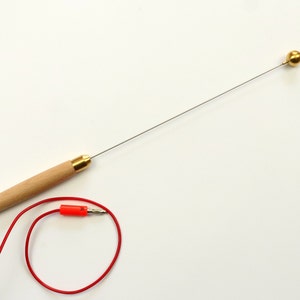 Puede incluir: Una herramienta con mango de madera, un alambre metálico delgado y una bola dorada en el extremo. También se muestra un cable rojo con un conector.