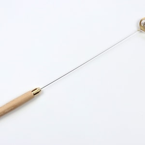 Puede incluir: Una herramienta larga y delgada con mango de madera, una varilla de metal y una pieza circular de latón en el extremo. La pieza de latón tiene una pequeña esfera de vidrio transparente en su interior.