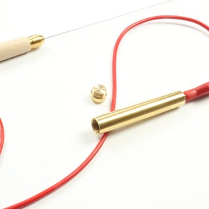 Puede incluir: Una herramienta de latón con un cable rojo conectado a un extremo y un mango de madera en el otro. La herramienta está abierta y el cable está enrollado alrededor de ella. Hay una pequeña tapa de latón en la mesa junto a la herramienta.