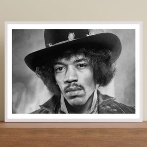 May include: A black and white photographic portrait of Jimi Hendrix wearing a wide-brimmed hat with a decorative band. The image captures a close-up view of the musician with his signature curly hair, mustache, and a dark jacket, presented in a frame.