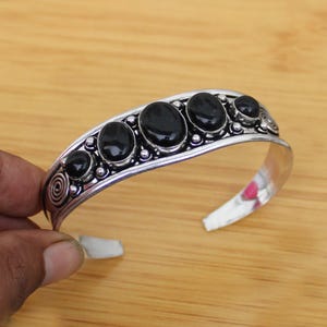 Könnte beinhalten: Silberfarbenes Armreif-Armband mit sechs ovalen und runden schwarzen Edelsteinen. Das Armband hat einen dekorativen Rand und ein Spiraldesign an einem Ende. Das offene Design ermöglicht einfaches Tragen.