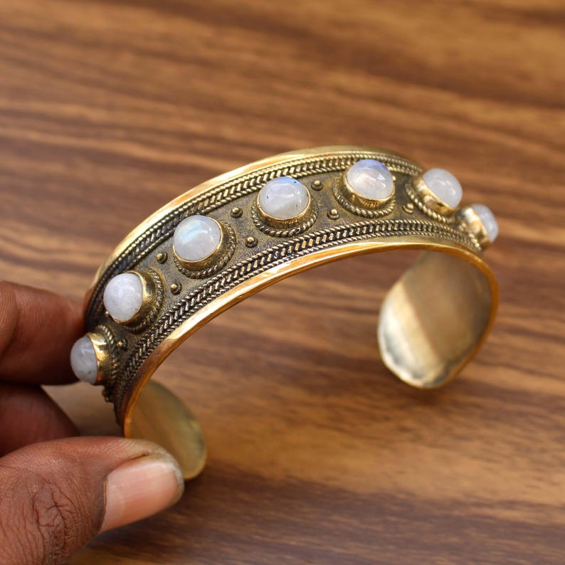 May include: A gold-toned cuff bracelet with a row of round, iridescent gemstones. The bracelet has a detailed, textured design with a vintage aesthetic. The gemstones are set in individual bezels, adding to the bracelet's ornate appearance.