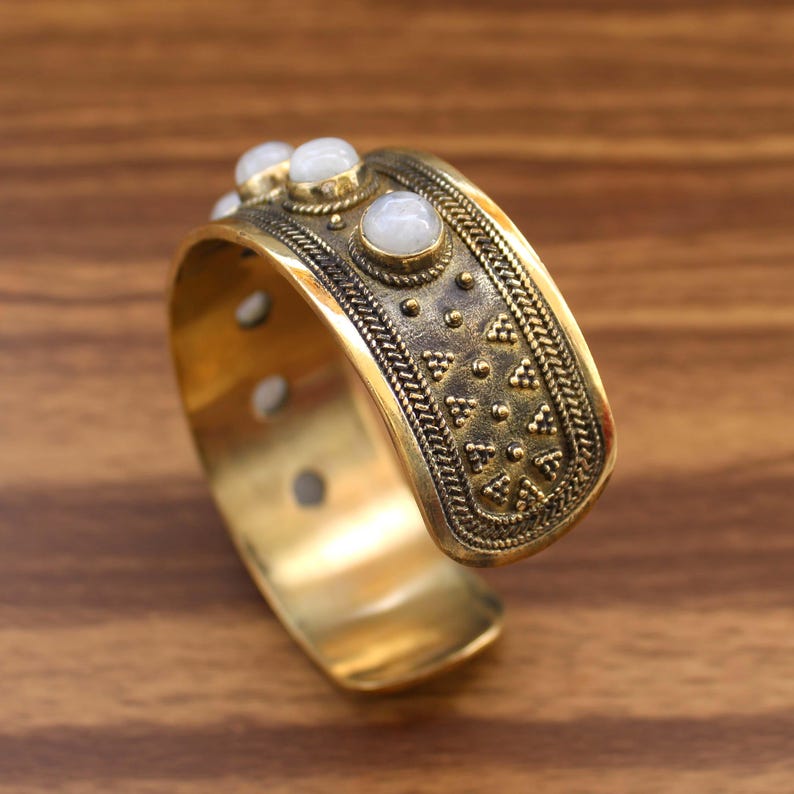 May include: A wide, gold-toned cuff bracelet with an ornate design. The bracelet features a textured surface with small, raised dots and triangular patterns. Three white, round stones are set along the top edge. The bracelet is displayed on a wooden surface.
