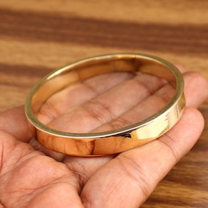 Puede incluir: Un brazalete de color dorado con una superficie plana y pulida. El brazalete es circular y parece estar hecho de metal. El brazalete se sostiene en la mano de una persona, mostrando su diseño y color.