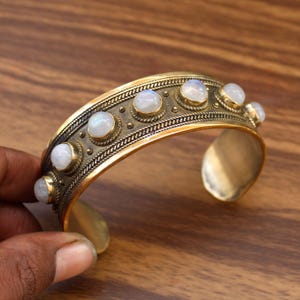 May include: A gold-toned cuff bracelet with a row of round, white gemstones. The bracelet has a textured design along the top edge and is open at the bottom. The gemstones are set in individual bezels.