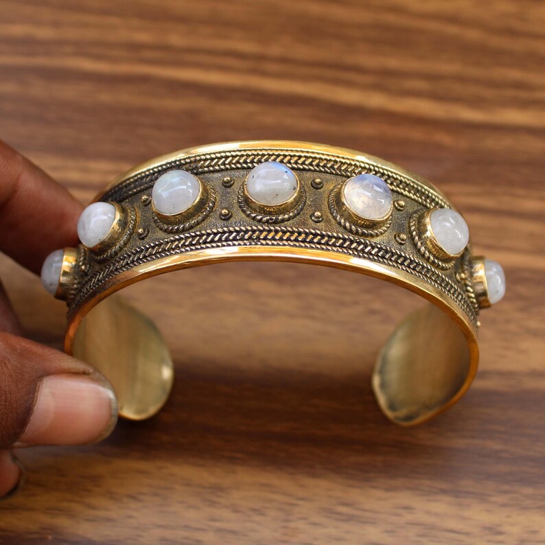 May include: A gold-toned cuff bracelet with six round, white gemstones. The bracelet has a detailed, textured design around the gemstones. The cuff is open at the bottom and is held by a hand. Jewelry.