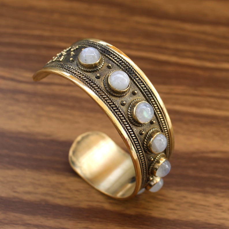 May include: A gold-toned cuff bracelet with a series of round, white gemstones. The bracelet features intricate detailing, including a braided pattern and small circular accents. The open design allows for easy wear.