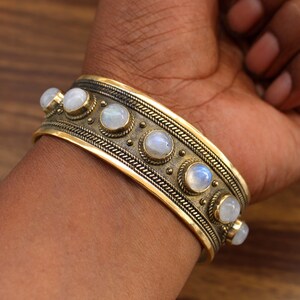 May include: A wide, gold-toned cuff bracelet with seven round, iridescent moonstone cabochons. The bracelet has an intricate, textured design with a braided pattern along the edges and small dot details. The bracelet is worn on a wrist.