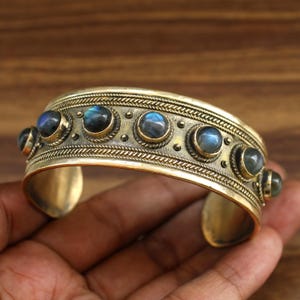 May include: An antique gold-coloured cuff bracelet. The bracelet features a series of round, iridescent blue-grey gemstones set in individual bezels. The cuff has a detailed, textured design with a braided pattern along the top and bottom edges.