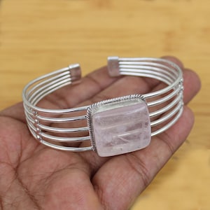 Könnte beinhalten: Silberfarbenes Armreif-Armband mit einem quadratischen Rosenquarzstein. Das Armband hat ein mehrsträngiges Design mit einem zentralen Steinfassungsbereich. Der Rosenquarz ist zartrosa.
