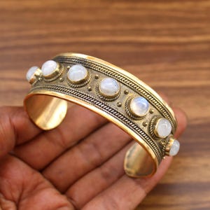 May include: A gold-toned cuff bracelet with a series of round, iridescent moonstones. The bracelet has an ornate design with a textured band and decorative edging. The open cuff design allows for easy wear.