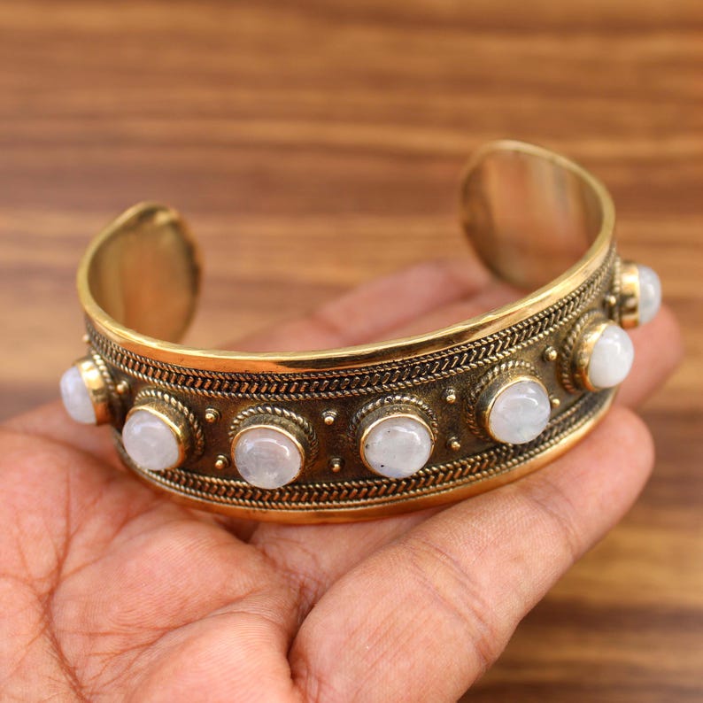 May include: A gold-toned cuff bracelet with a row of six round, white gemstones. The bracelet has an open design and is decorated with intricate patterns along the edges. A piece of jewelry.