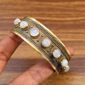 May include: A gold-toned cuff bracelet with a series of round, white moonstone cabochons. The bracelet features intricate detailing and a textured design. The bracelet is held by a hand.