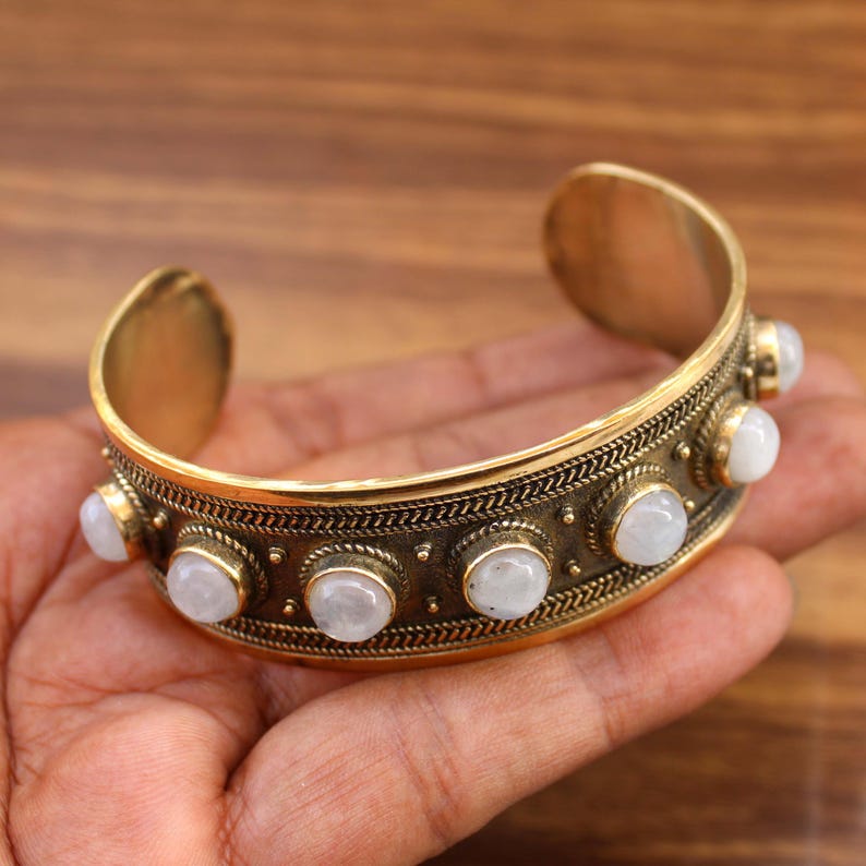 May include: A gold-toned cuff bracelet with a decorative band featuring small, round, white gemstones. The bracelet has an open design and is held in a hand, showcasing its intricate details and craftsmanship.