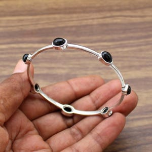 May include: A silver bangle bracelet featuring five evenly spaced, smooth, round black stones set in individual metal bezels. The bracelet has a thin, polished silver band that connects each of the stone settings in a continuous circular design.