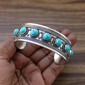 May include: A silver cuff bracelet with turquoise stones. The bracelet features a detailed design with a row of turquoise cabochons set in individual bezels. The metalwork includes a braided pattern along the edges and small circular accents.