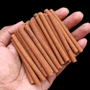 Puede incluir: Una colección de palitos de incienso cilíndricos marrones, sostenidos en una mano sobre un fondo negro. Los palitos de incienso son de tamaño y color uniformes, con una textura ligeramente rugosa.