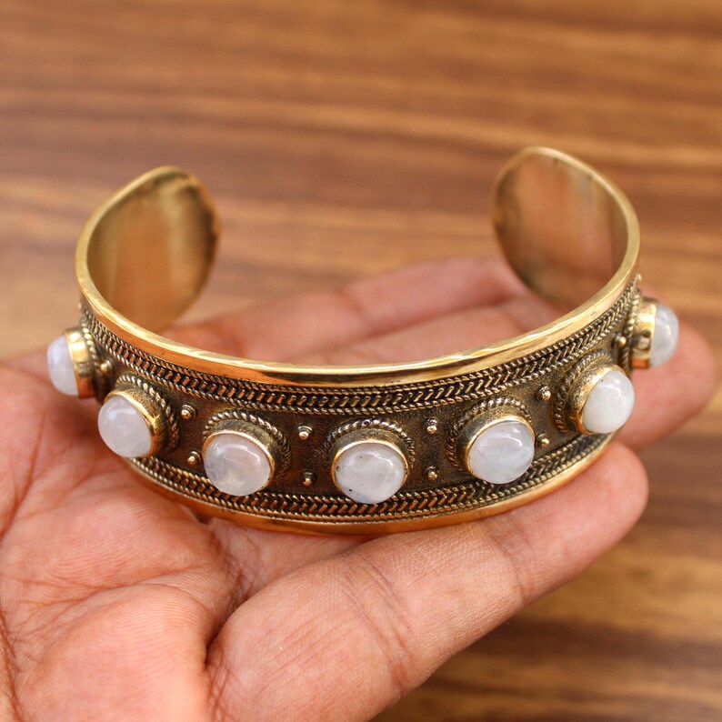 May include: A gold-toned cuff bracelet with a decorative band featuring a series of white, round gemstones. The bracelet has an open design and is held in a hand, showcasing its intricate details and craftsmanship. The bracelet is a piece of jewelry.