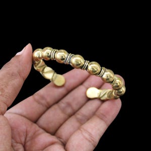 Puede incluir: Una pulsera de puño dorada con una serie de cuentas redondas. La pulsera tiene un diseño abierto con terminales redondeados. Las cuentas están conectadas por finas bandas de color plateado. La pulsera se sostiene sobre un fondo negro.