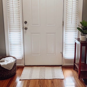 May include: A white door with a white doormat in front of it. The door has a single pane of glass and a set of blinds on either side. The doormat is made of a woven material and has a light brown color.