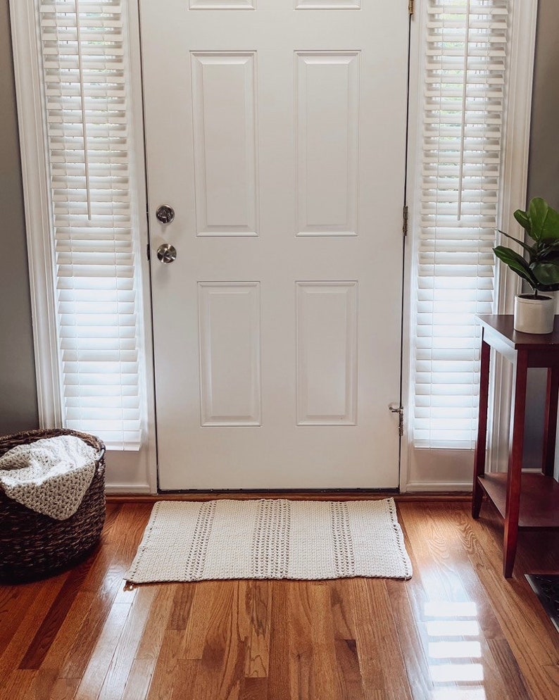 May include: A white door with a white doormat in front of it. The door has a single pane of glass and a set of blinds on either side. The doormat is made of a woven material and has a light brown color.