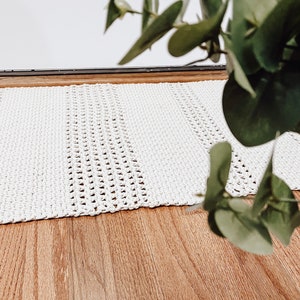 May include: A white crocheted doormat with a textured design. The doormat is placed on a wooden floor.
