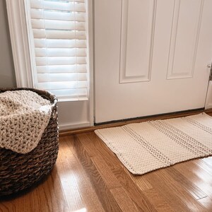 May include: A woven basket with a cream-colored crocheted blanket inside, sitting on a wooden floor next to a white door and a cream-colored crocheted rug.
