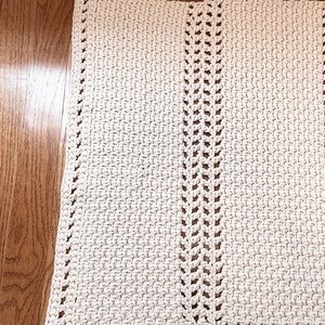 May include: A white crocheted rug with a simple design featuring two rows of open stitches down the center. The rug is laid out on a wooden floor.