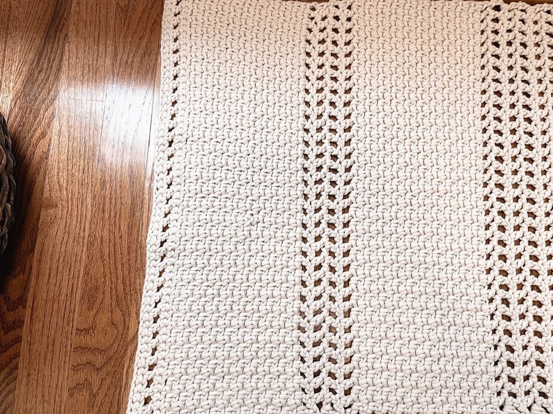 May include: A white crocheted rug with a simple design featuring two rows of open stitches down the center. The rug is laid out on a wooden floor.