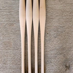 May include: Four unfinished wooden baseball bats. The bats are light-colored, with a smooth, natural wood grain. Each bat has a rounded handle and a tapered barrel. The bats are arranged vertically against a textured surface.
