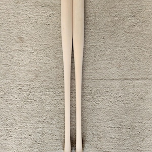 May include: Two unfinished wooden baseball bats, standing vertically against a textured, neutral-toned background. The bats are light in color, with a smooth, cylindrical handle and a wider barrel end. The bats are ready for customization.