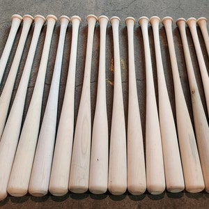 May include: A row of 18 unfinished wooden baseball bats laid out on a concrete floor.