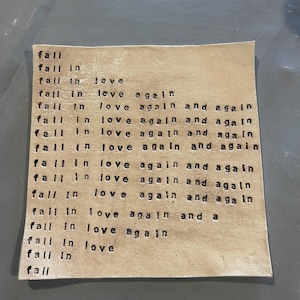 May include: A beige canvas with the phrase "fall in love again and again" repeated multiple times in black lettering.