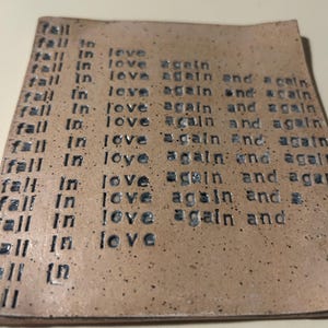 May include: A square, tan ceramic coaster with the phrase "fall in love again and again" stamped repeatedly in a dark grey font. The coaster has a speckled texture and a slightly uneven surface.