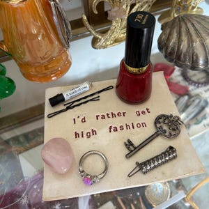 Puede incluir: Una colección de accesorios que incluye un frasco de esmalte de uñas rojo, un anillo con una piedra rosa, una llave decorativa y un pequeño vial de perfume. Los artículos están dispuestos sobre una superficie cuadrada con el texto "I'd rather get high fashion".