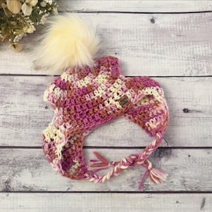 May include: A pink, white, and yellow crocheted hat with a fluffy white pom pom. The hat has a long braided strap.
